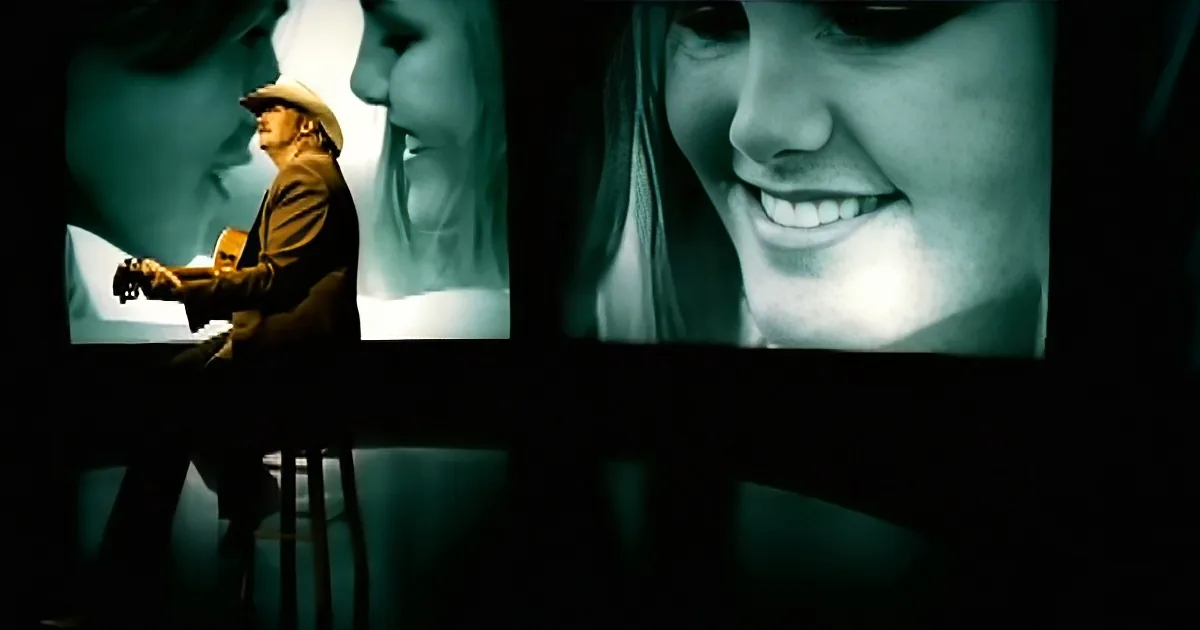A man in a cowboy hat plays guitar on a stool, surrounded by large screens showing smiling faces.
