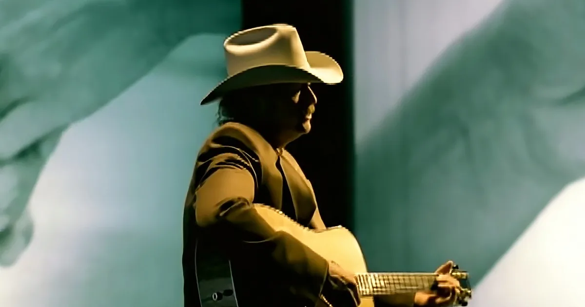 A man in a cowboy hat plays an acoustic guitar against a teal backdrop.
