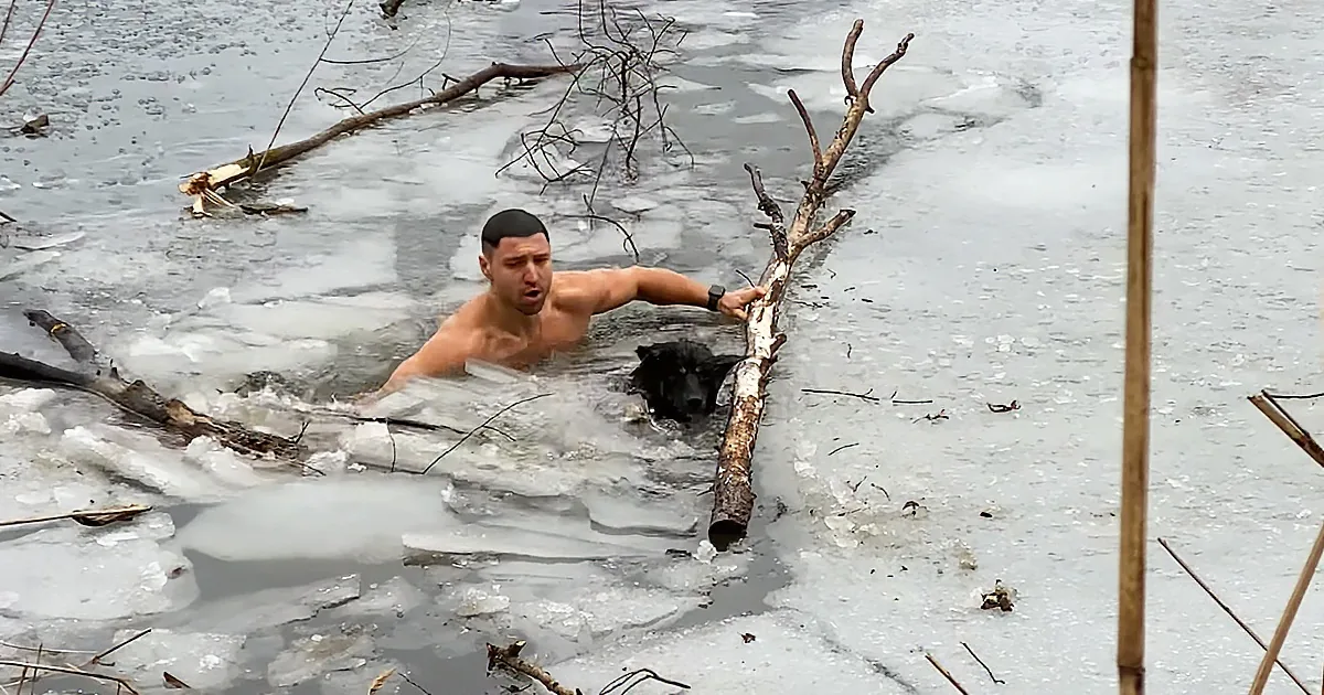 A shirtless man struggles in icy water, holding onto a log amidst broken ice.