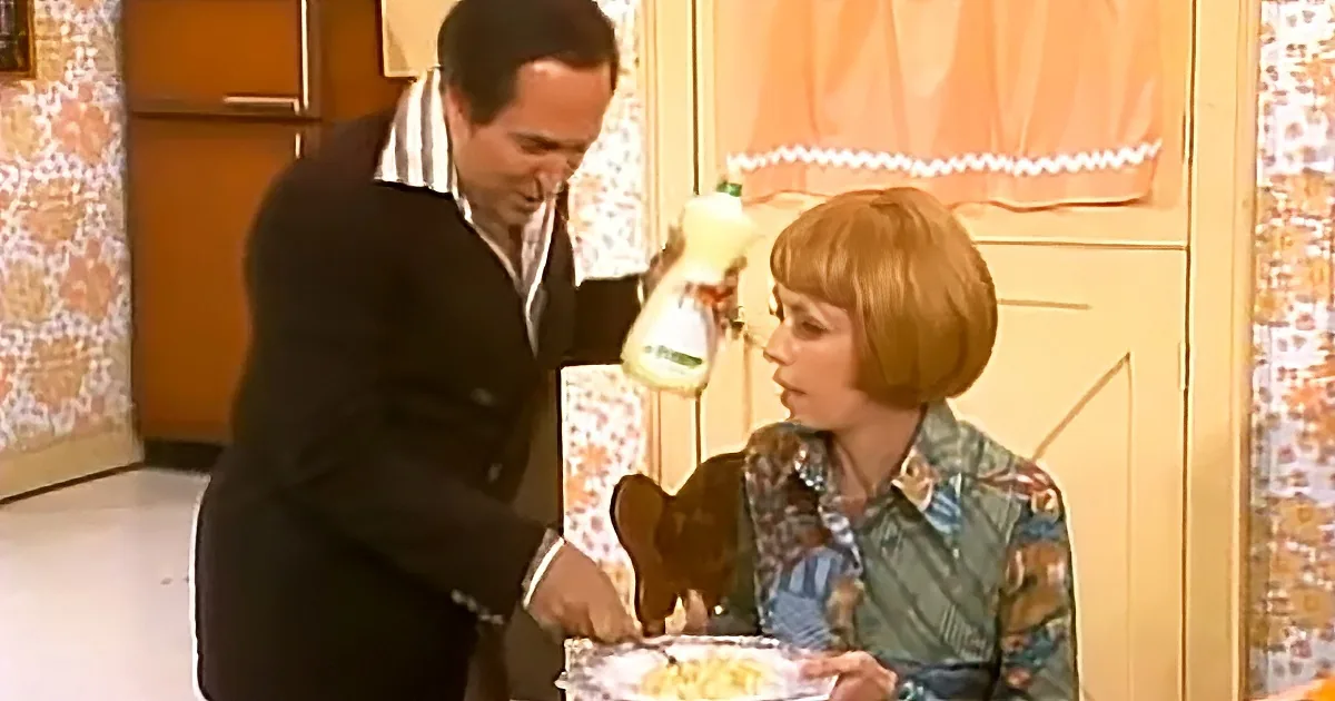 A man in a suit pours milk into a glass while a woman in a floral shirt looks on in a kitchen.