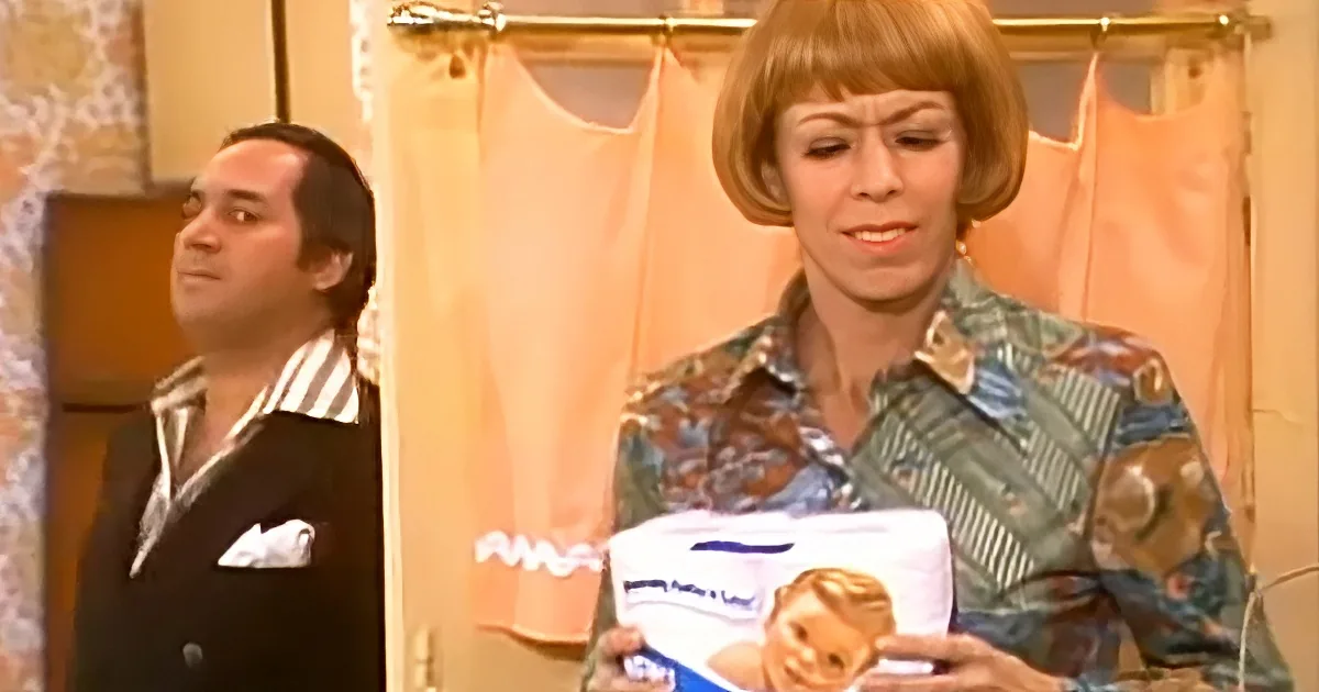 A woman holds a box of feminine hygiene products, while a man in a suit looks on in a bathroom.