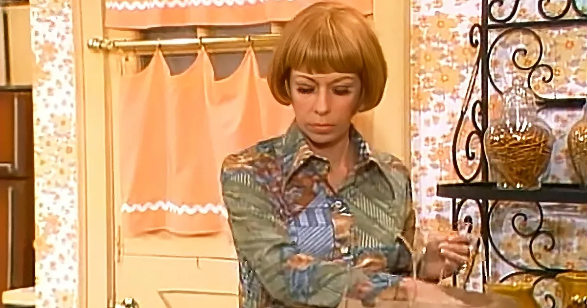A woman with short hair stands in a kitchen, holding a glass, wearing a patterned shirt.