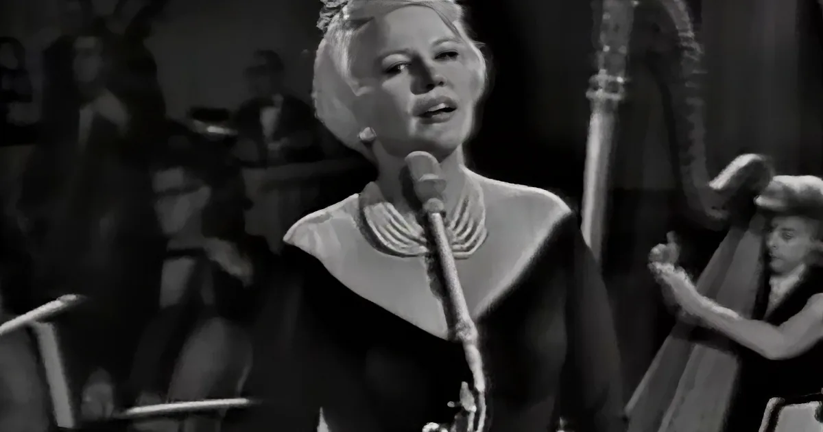 A woman in a black dress sings into a microphone, surrounded by a band and audience.