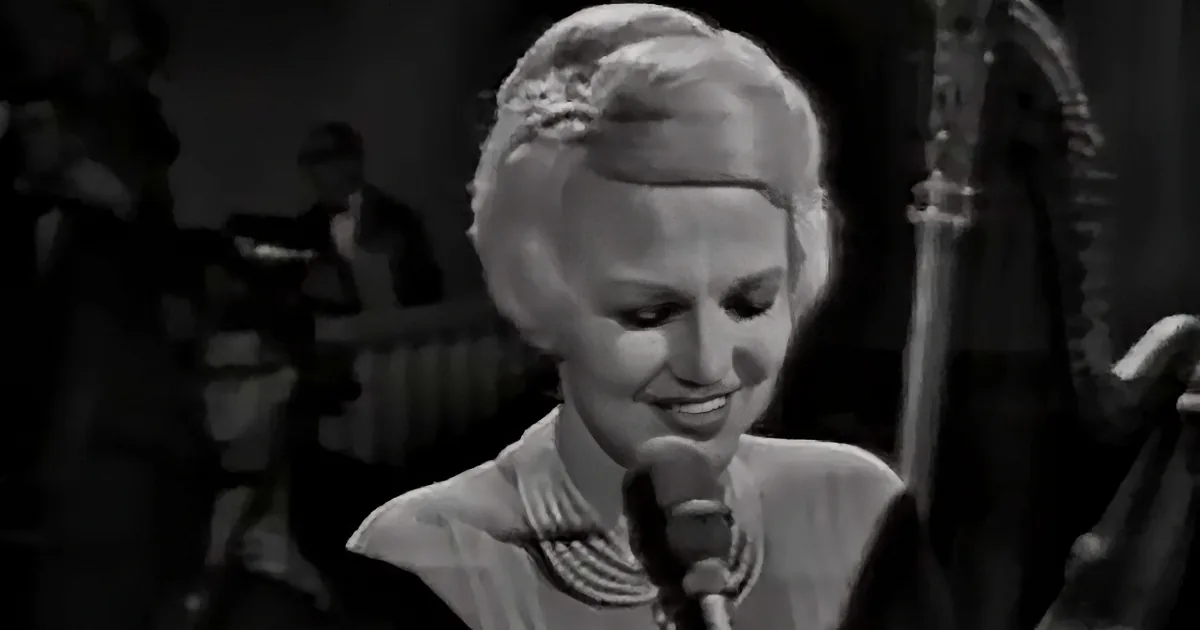 A woman with blonde hair, wearing a striped dress, smiles while singing into a microphone.