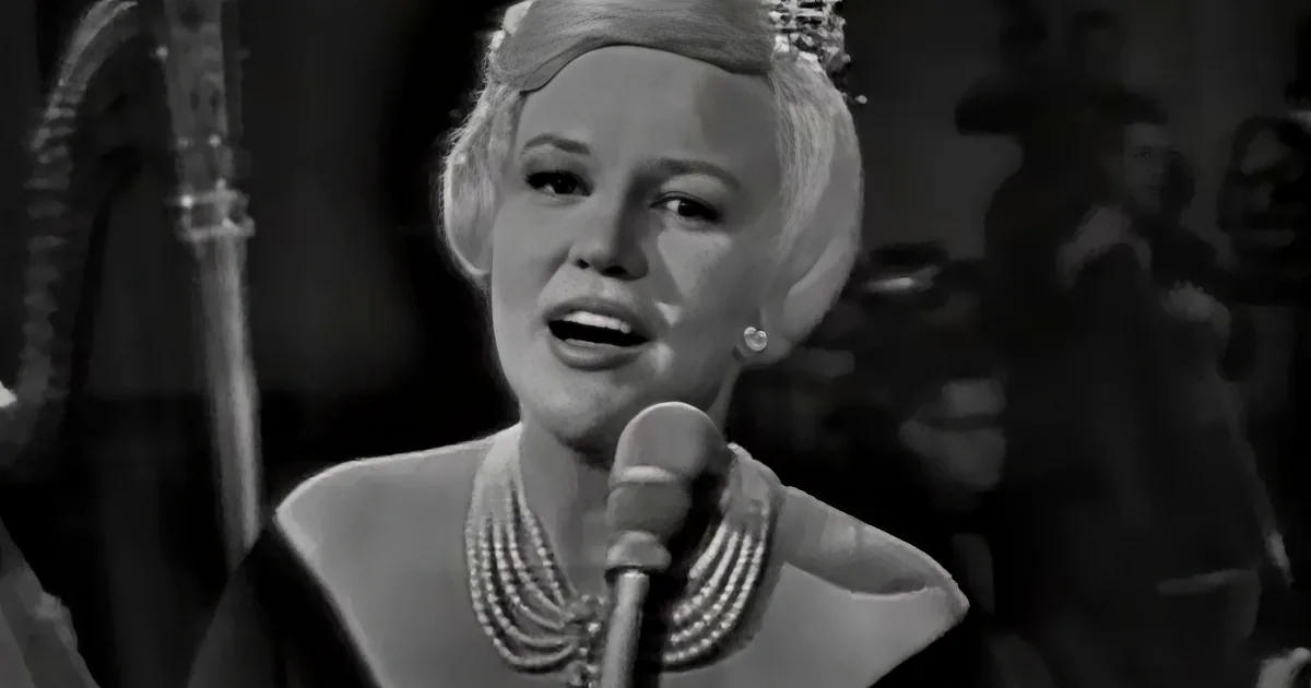 A woman sings into a microphone, wearing pearls and a tiara, with a band in the background.