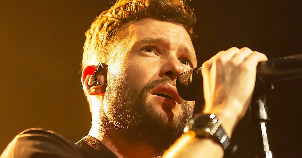 A man with a beard and earpiece sings into a microphone under warm stage lighting.