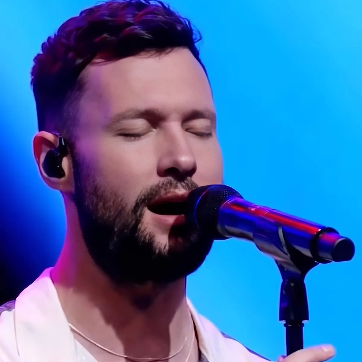 A man with a beard and earpiece sings into a microphone on stage with a blue background.