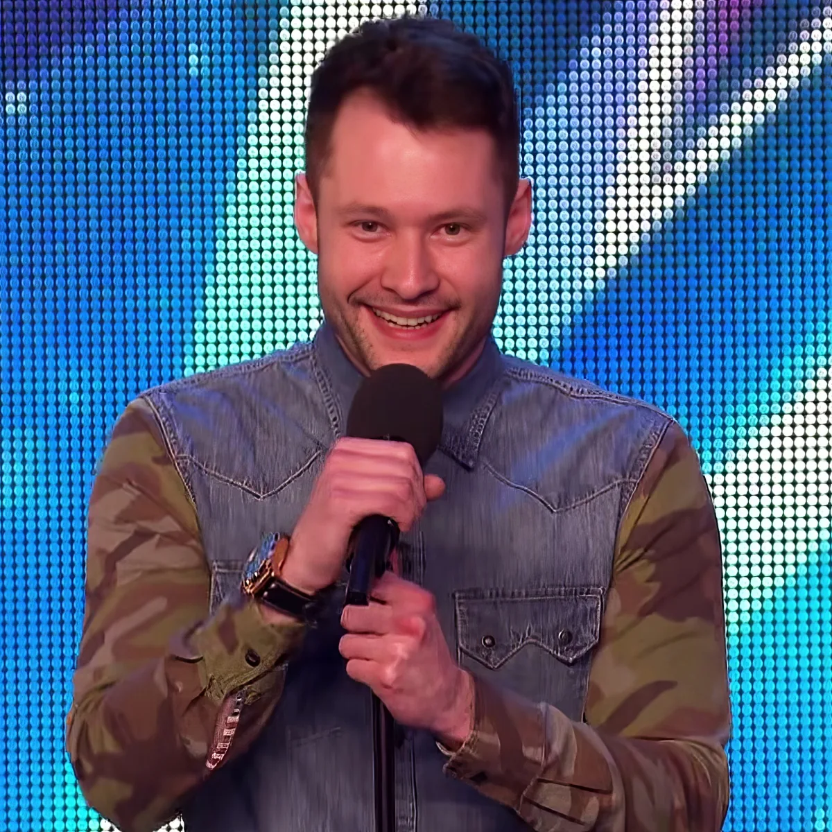 A man in a camouflage jacket and denim vest holds a microphone, smiling against a blue LED background.