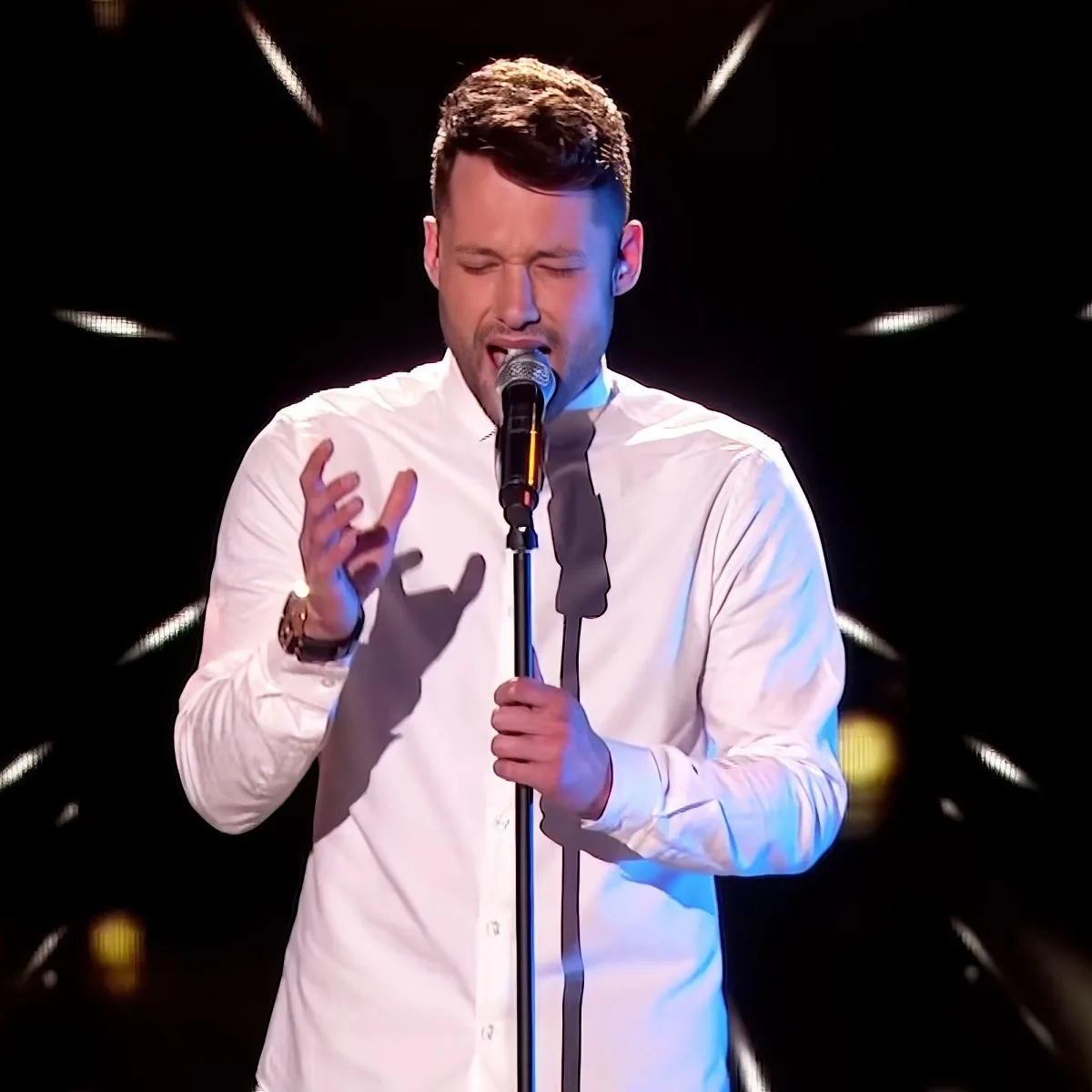 A man in a white shirt sings passionately into a microphone, gesturing with his hands.