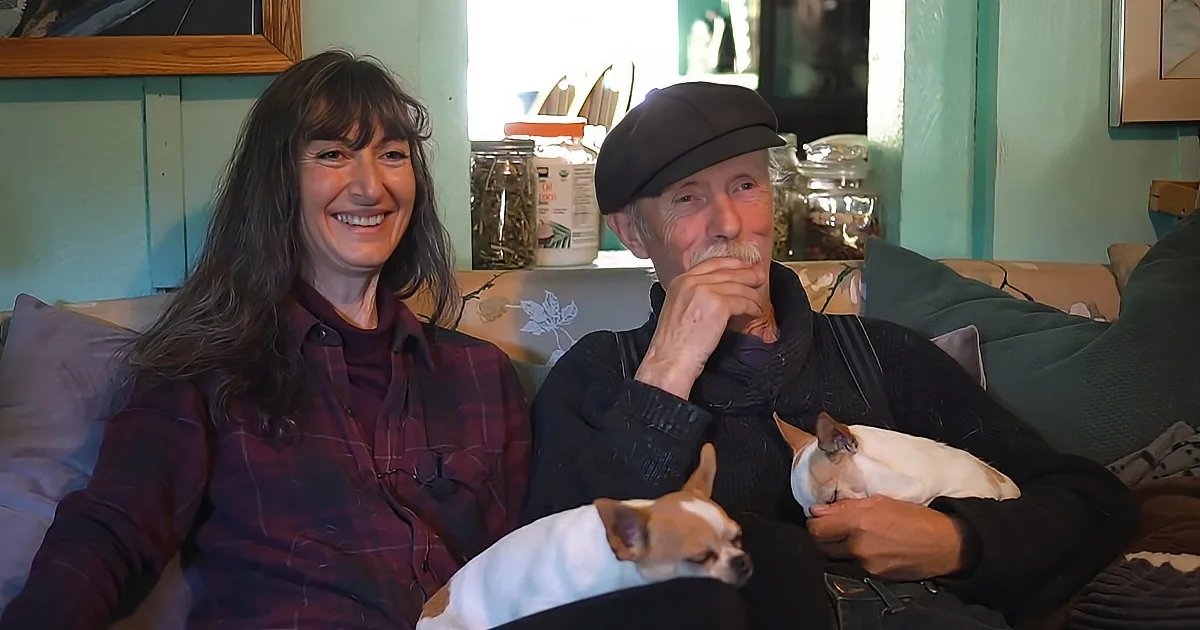 A couple sits on a couch with two small dogs, smiling and relaxed in a cozy room.