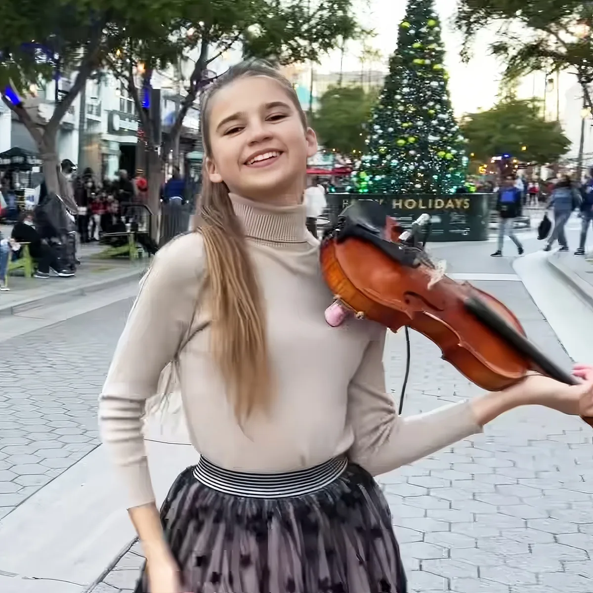 A girl smiles while holding a violin in a festive outdoor setting with a Christmas tree and holiday decorations.