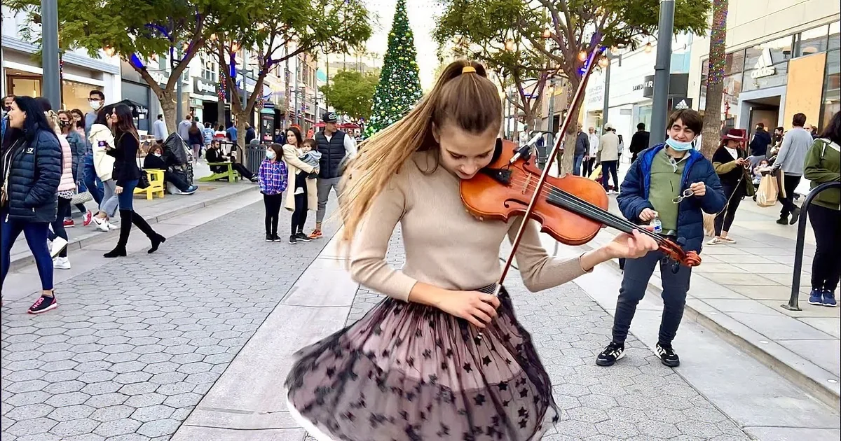 A girl smiles while playing the violin on a city street with holiday decorations and passersby.