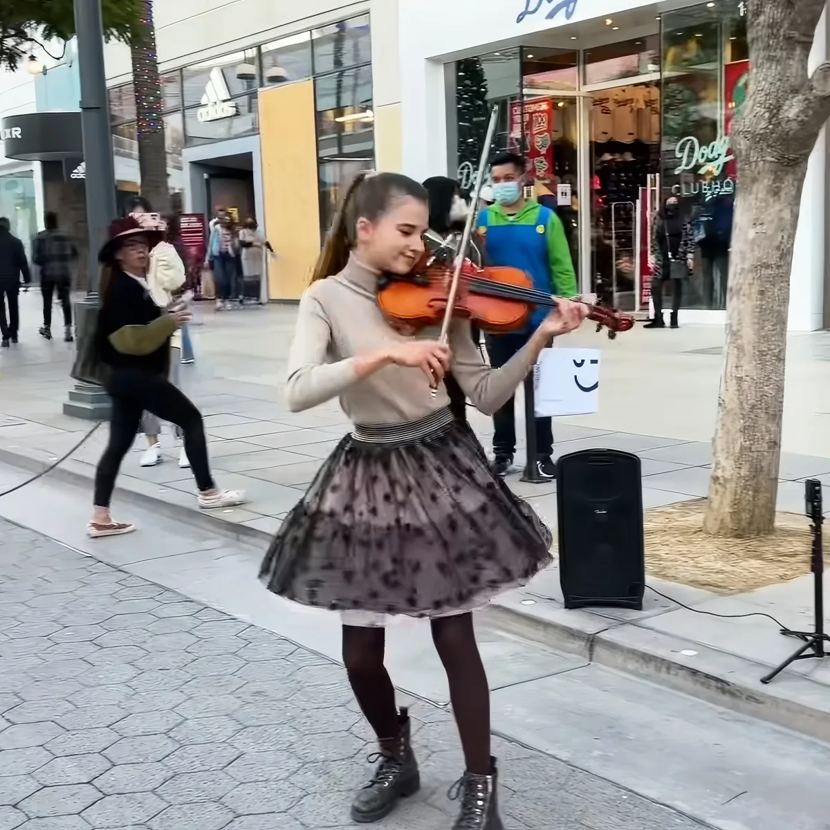 A young girl in a skirt and turtleneck plays the violin on a city sidewalk, surrounded by passersby and storefronts.