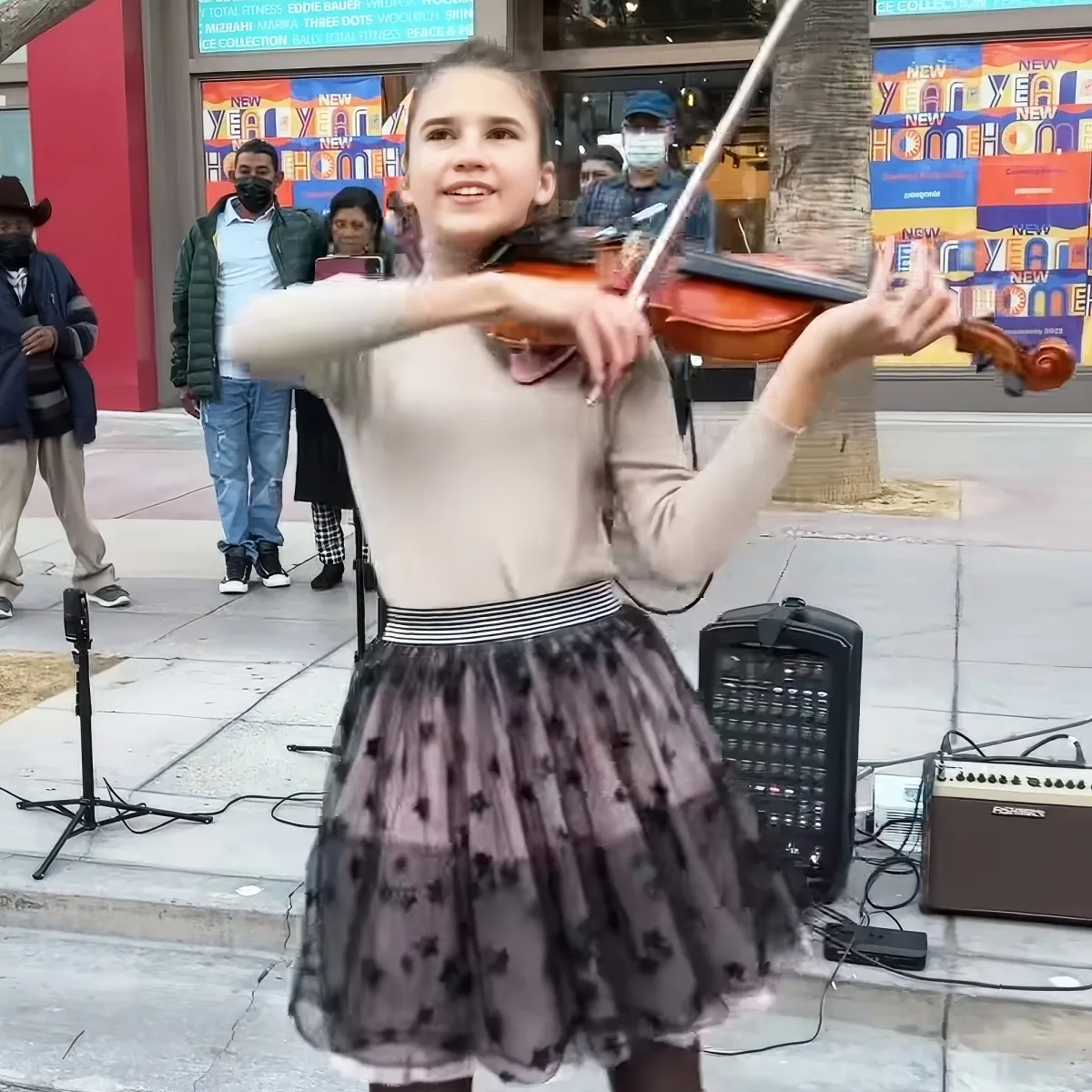 A young girl in a tutu plays the violin on a sidewalk, surrounded by onlookers and street performers.