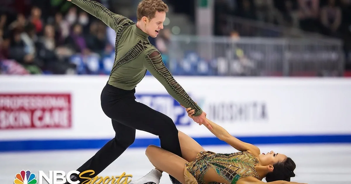 A figure skater in a gold outfit is being lifted by another skater in a green top on an ice rink.
