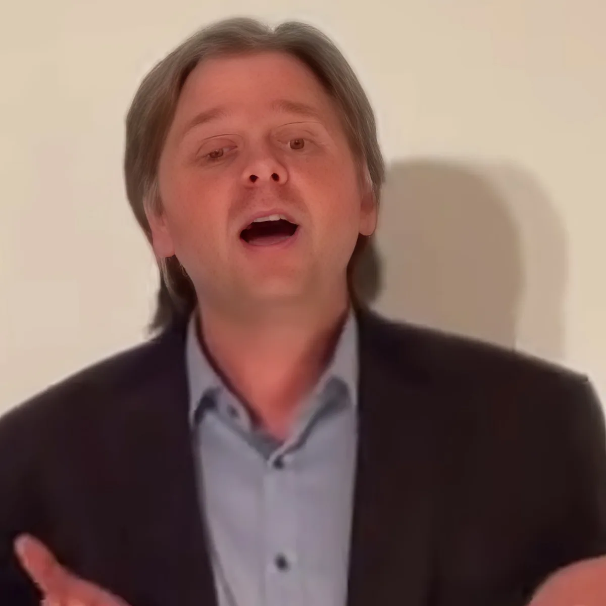A man in a suit and shirt appears to be speaking or singing with his mouth open.