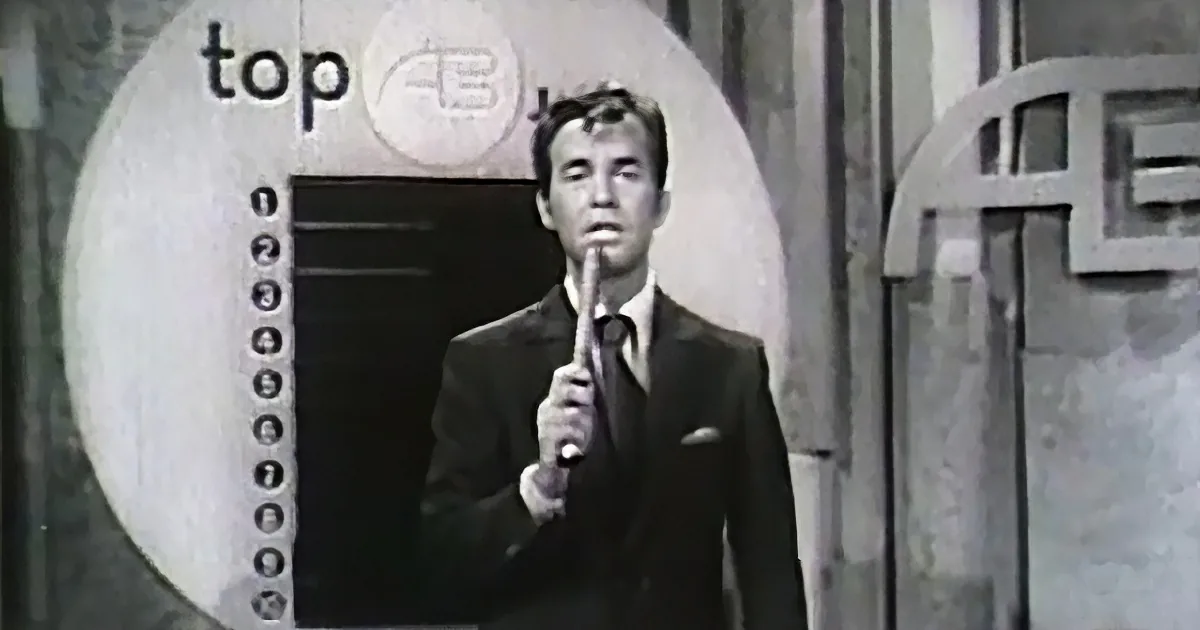 A man in a suit holds a microphone, standing in front of a "Top 40" sign with a countdown list.