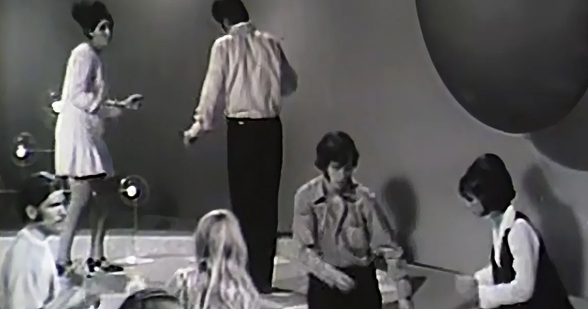 A person in a white dress walks on stage, while others in formal attire stand and sit nearby.