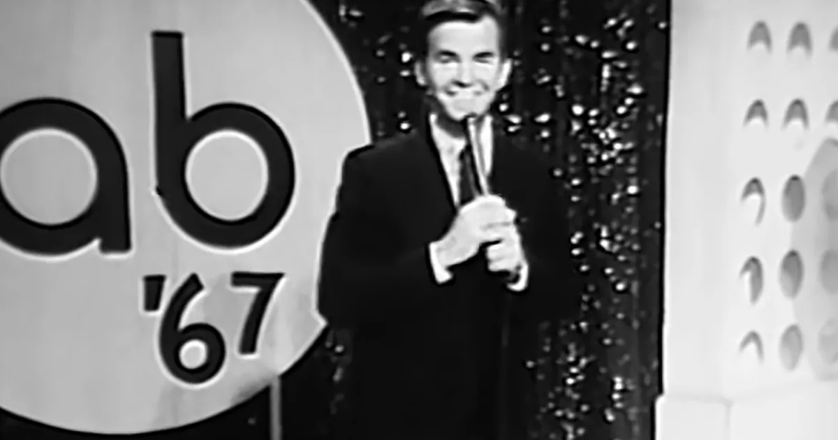 A man in a suit holds a microphone on stage with "abc '67" in the background.