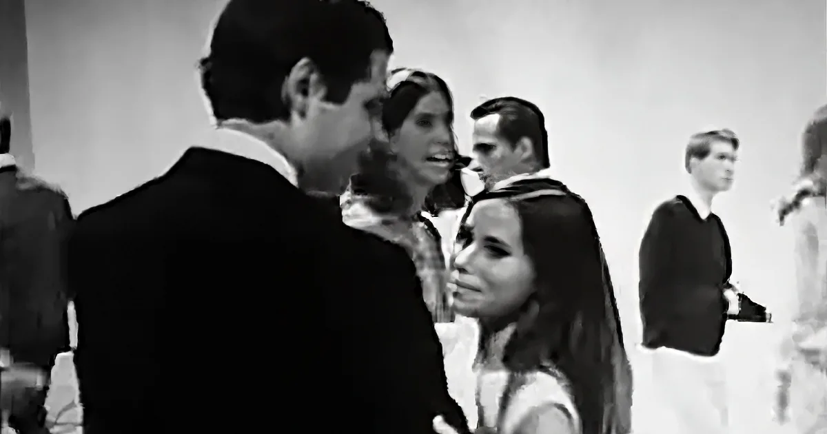A man in a suit is talking to a woman with long hair, surrounded by other people in a social setting.