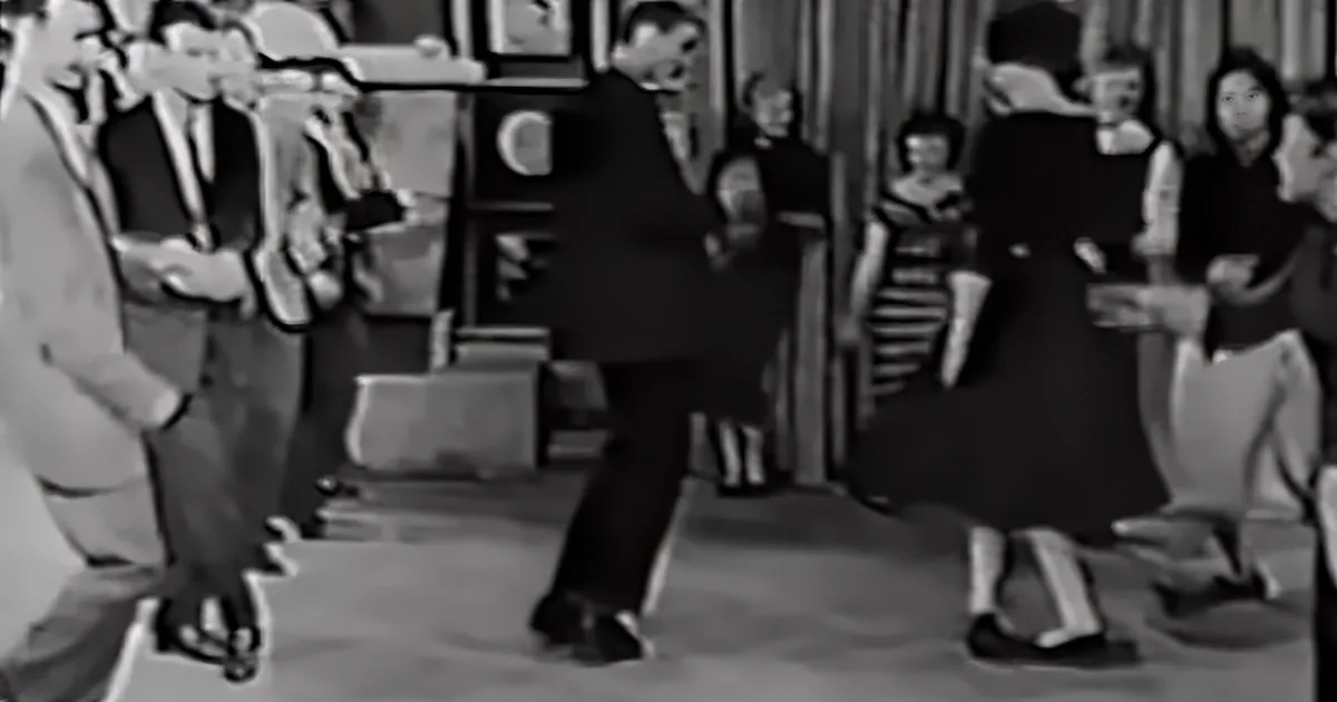 A man in a suit dances in a lively room with other people watching.
