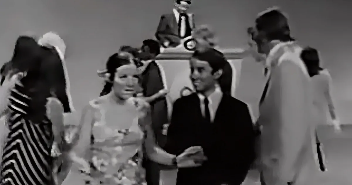 A man in a suit and tie is dancing with a woman in a dress, surrounded by other people in a social setting.