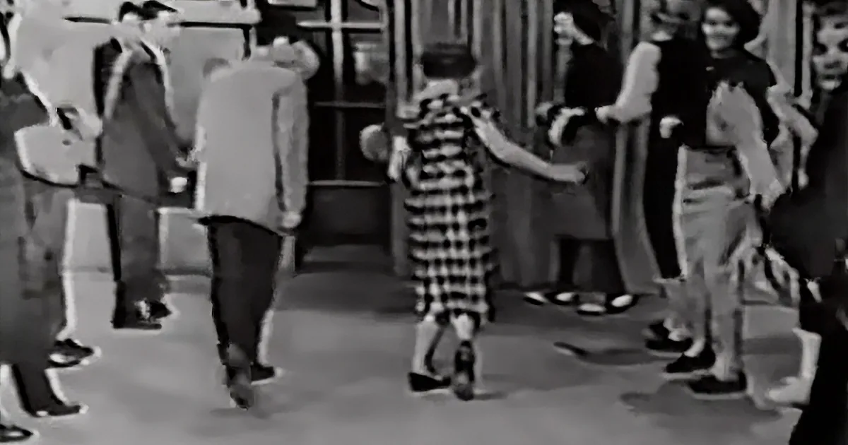 A young girl in a checkered dress dances amidst a crowd of people in a vintage setting.