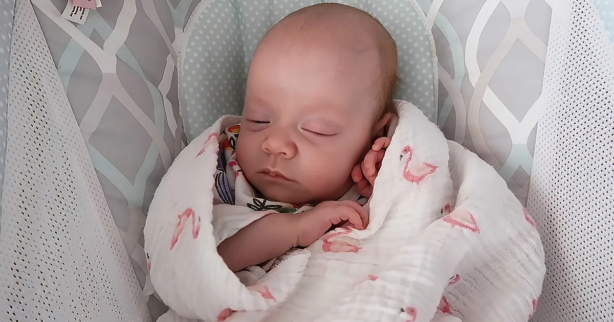 A newborn baby is sleeping peacefully in a white and pink swaddle, nestled in a patterned crib.