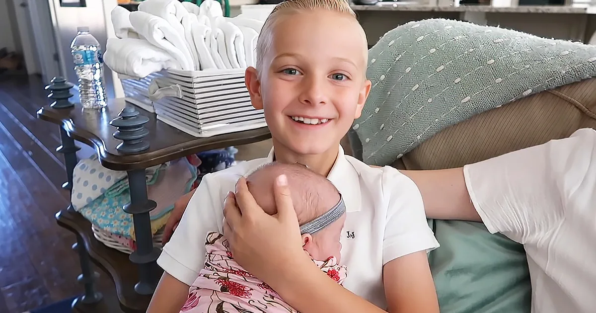 A young boy in a white shirt holds a newborn baby, smiling, with a water bottle and folded towels in the background.