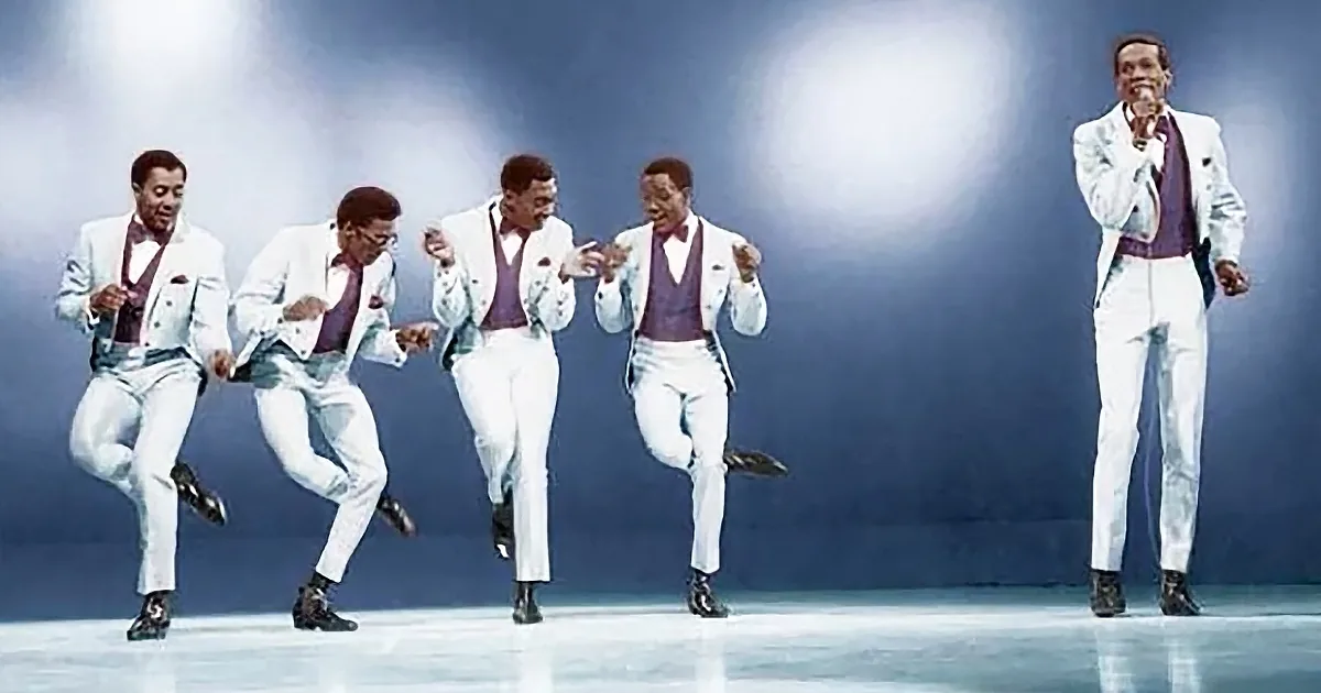A group of men in white suits and purple vests perform a synchronized dance routine on stage.