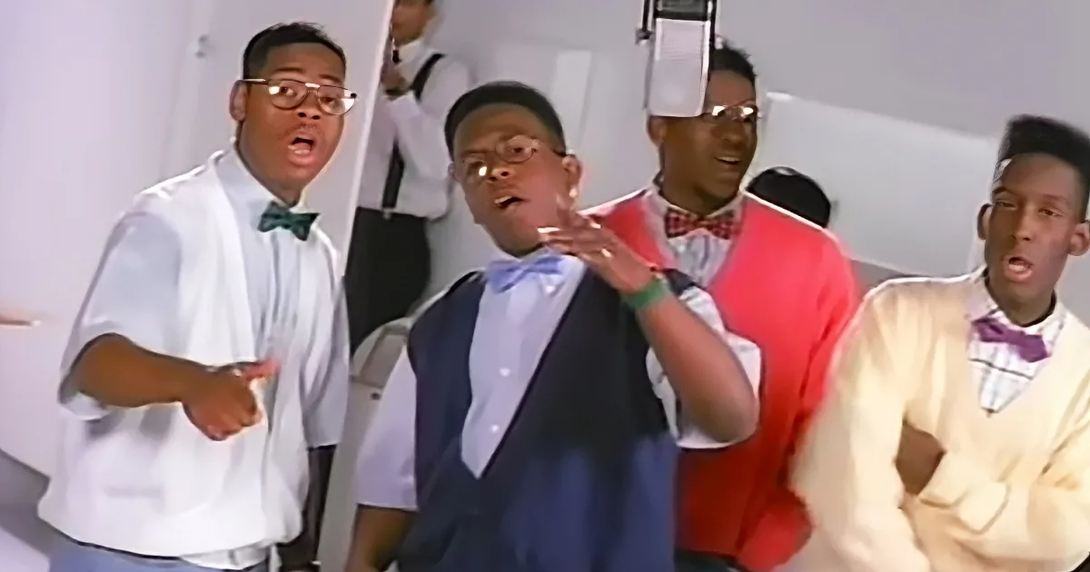 A man in a white shirt and bow tie is singing and gesturing, surrounded by other men in formal attire.