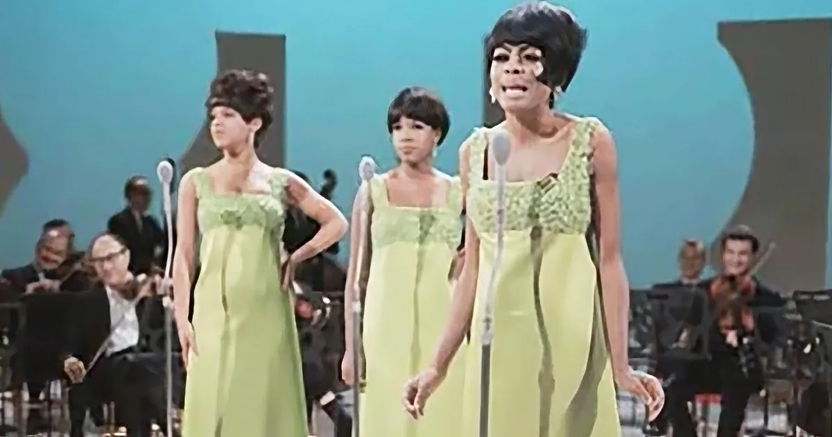 A woman in a green dress sings on stage with two backup singers, accompanied by an orchestra.