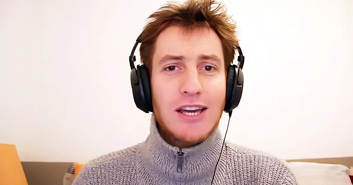 A person wearing headphones and a gray sweater is speaking in front of a plain background.