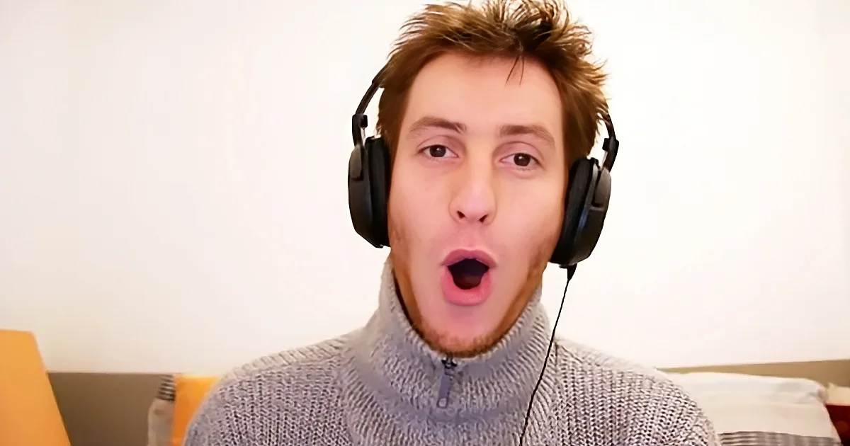 A person wearing headphones and a gray sweater is shown in a close-up, mouth open.