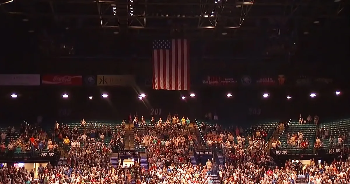 A large crowd fills a packed arena with an American flag hanging prominently above.