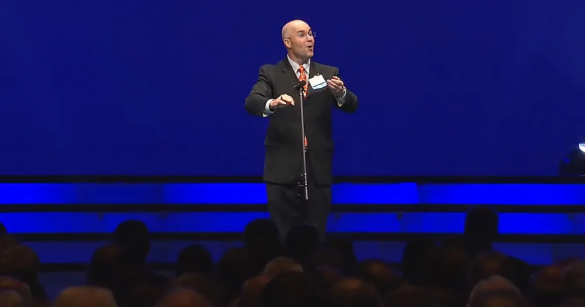 A man in a suit stands on stage, speaking to an audience with a microphone.