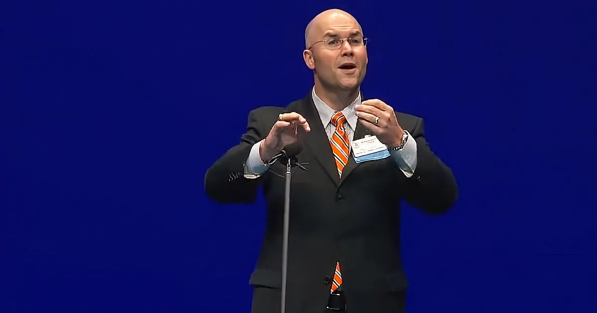 A man in a suit and tie is speaking at a podium with a blue background.