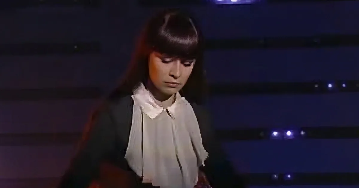 A person with long hair and a white shirt stands in a dimly lit room, looking down.