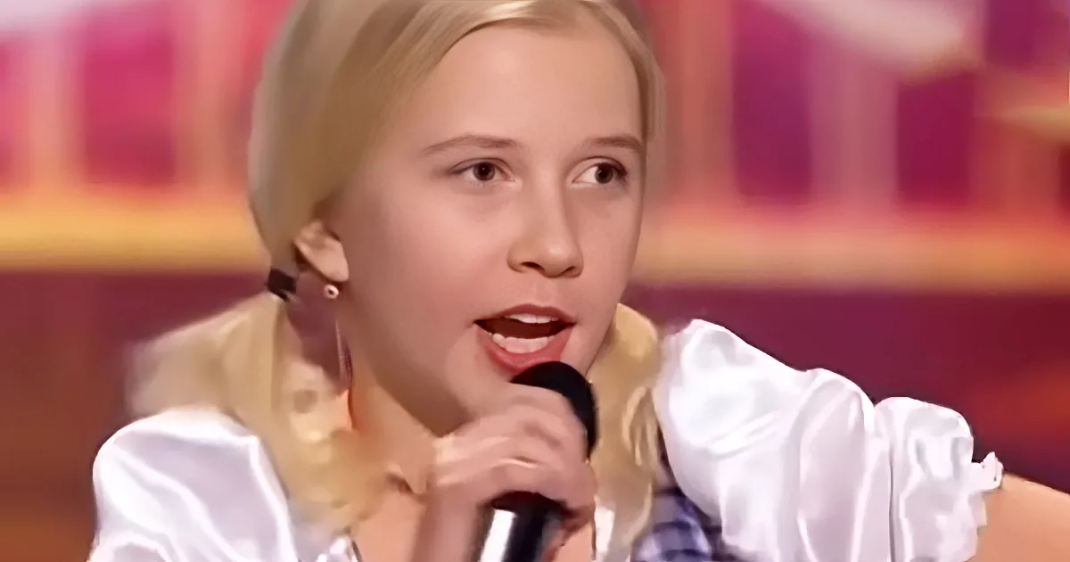 A young person with blonde hair, wearing a white outfit, sings into a microphone.