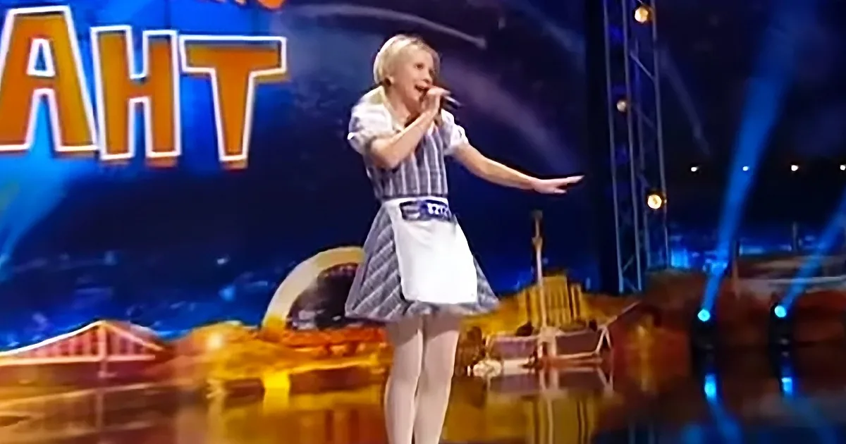A young person in a striped dress sings on stage with a microphone, illuminated by blue lights.