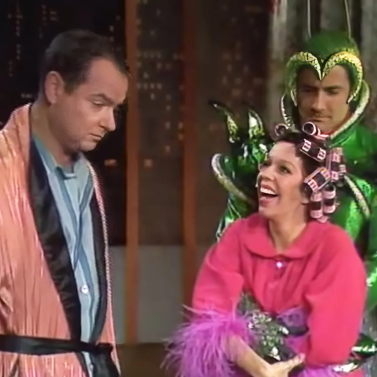 A man in a pink robe stands beside a woman in a pink coat, both looking at something off-screen, with a person in a green...