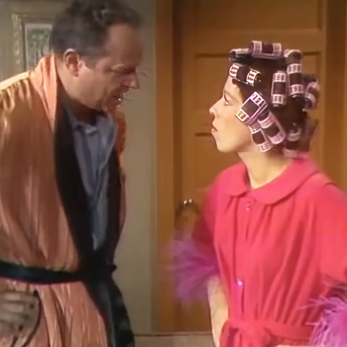 A man in a robe leans towards a woman in a pink dress with curlers in her hair.