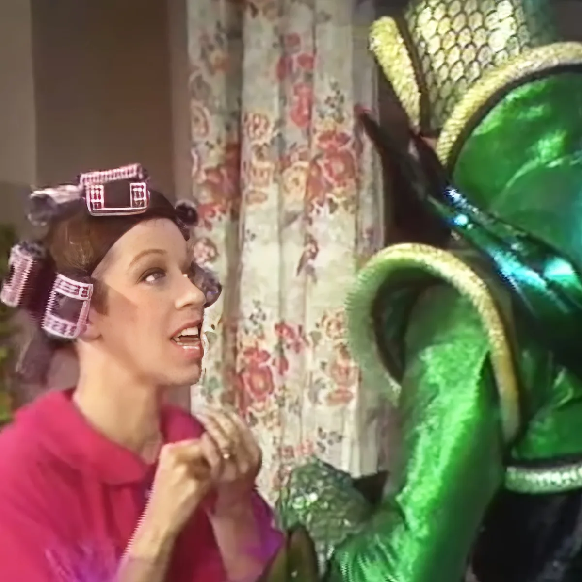 A woman with curlers in her hair looks surprised at a person in a green costume with a large headpiece.