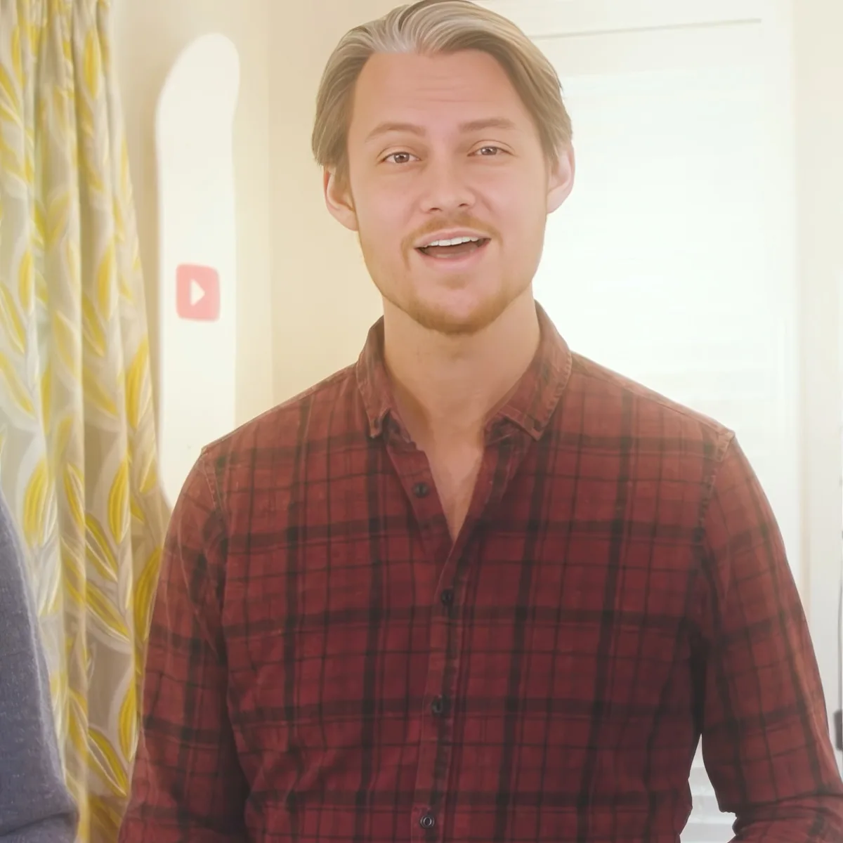 A man with a beard, wearing a red plaid shirt, stands indoors with a YouTube play button visible in the background.