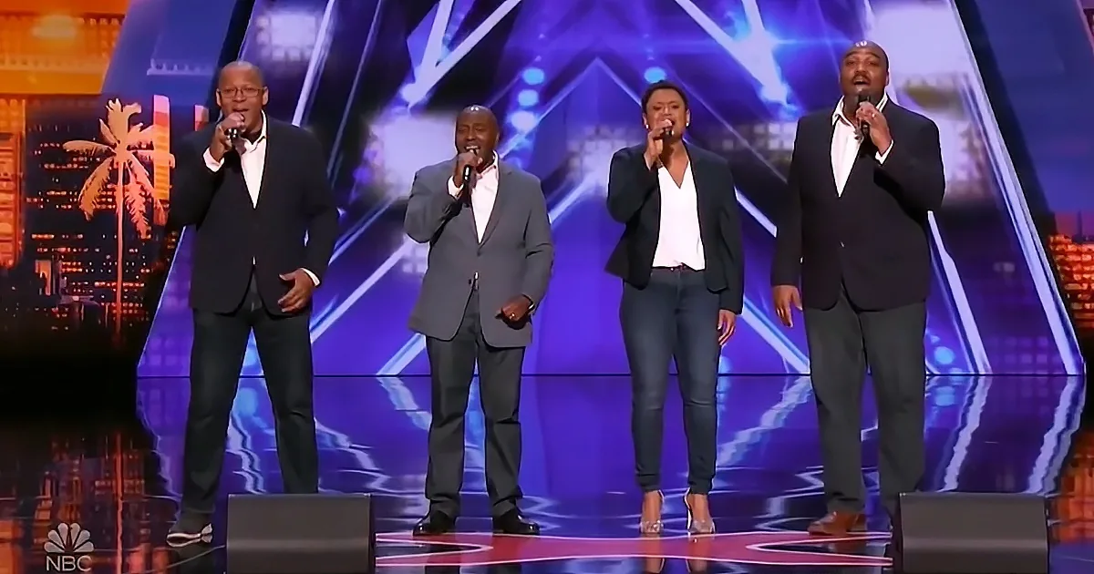 Four performers in formal attire sing on a stage with vibrant lighting and cityscape backdrop.