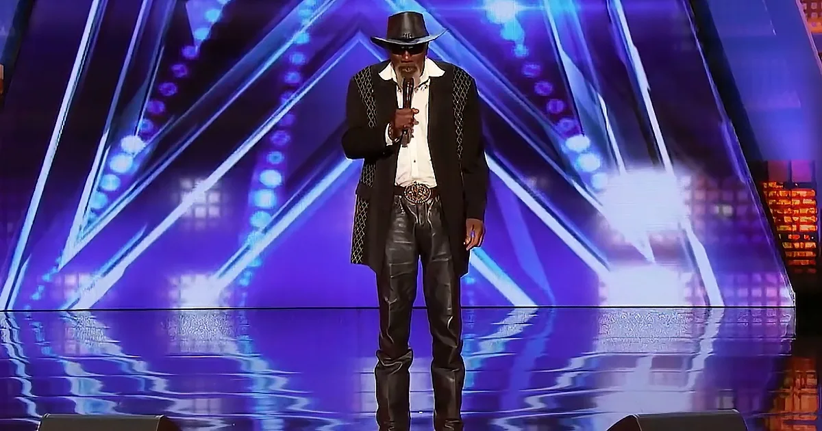 A man in a black suit, hat, and sunglasses stands on stage holding a microphone.
