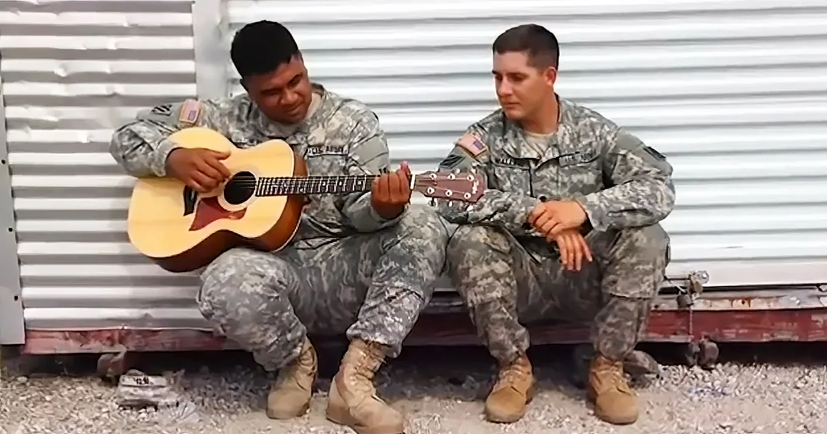 Two soldiers in camouflage uniforms sit on a bench, one playing a guitar while the other watches.