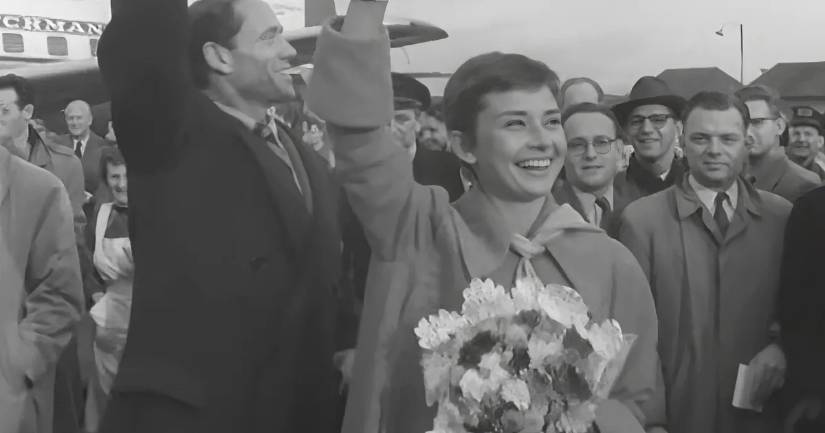 A smiling woman holds flowers, waving to a crowd, with a man in a suit beside her.