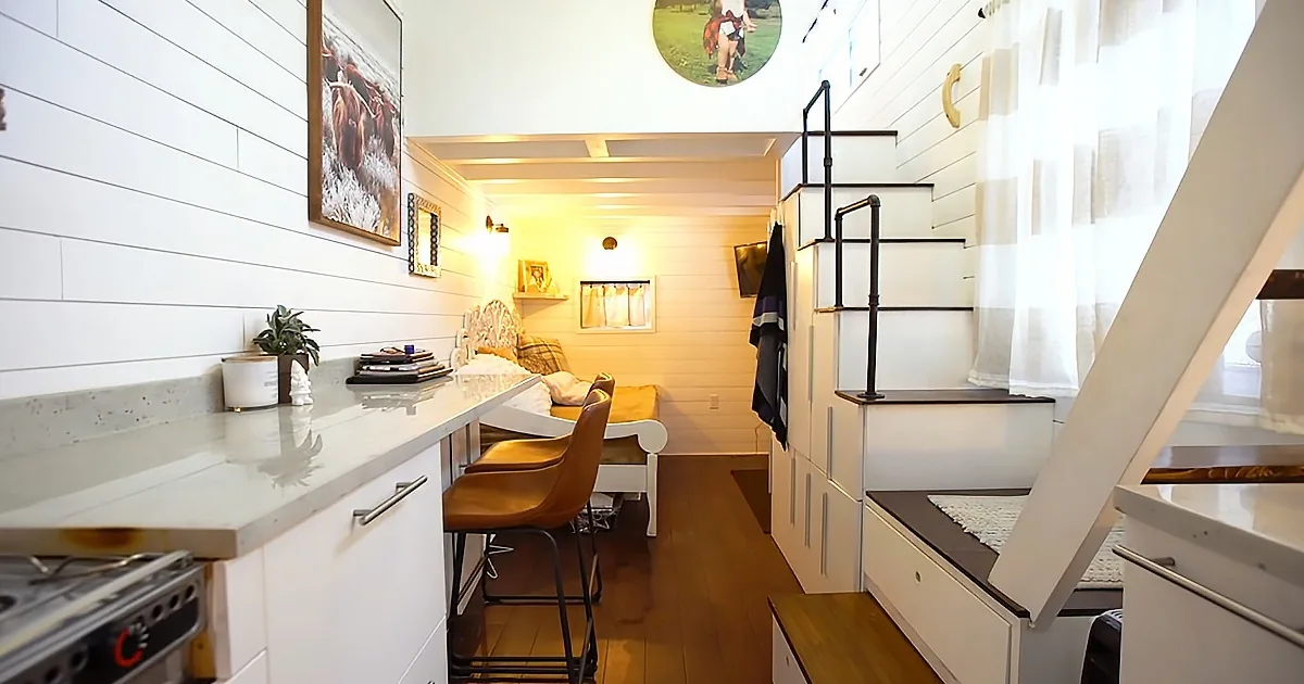 A cozy, compact living space with white walls, a desk, a bed, a staircase, and a kitchenette.