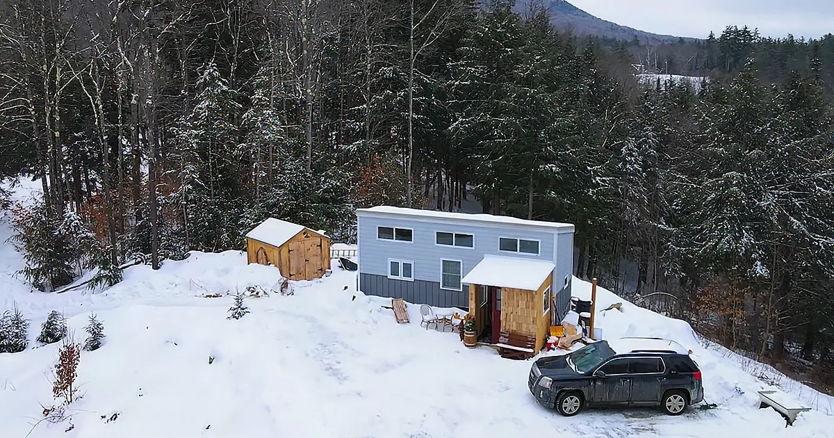 A small, snow-covered cabin with a car parked in front, surrounded by a snowy forest.